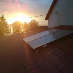 Panele słoneczne na dachu domu, widoczne krople deszczu. W tle zachodzące słońce i drzewa. Czerwona dachówka. Ujęcie z dołu, lekko z boku.