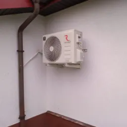 Kremowy klimatyzator Rotenso Fresh zamontowany na białej, chropowatej ścianie zewnętrznej budynku, widoczna brązowa rynna i fragment dachu.