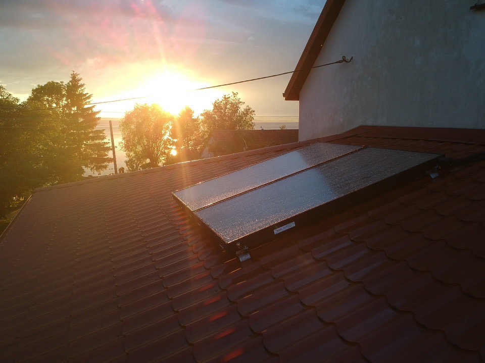 Panele słoneczne na dachu domu, widoczne krople deszczu. W tle zachodzące słońce i drzewa. Czerwona dachówka. Ujęcie z dołu, lekko z boku.