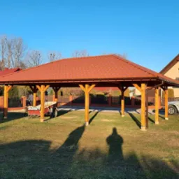 Nowoczesna, drewniana konstrukcja altany z czerwoną dachówką, widoczna na tle domu z balkonem i zaparkowanym samochodem. Na trawniku cień osoby robiącej zdjęcie.