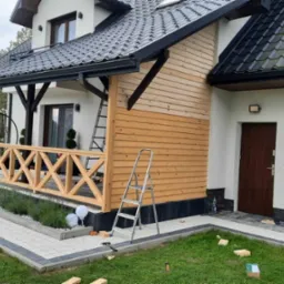 Dom jednorodzinny z drewnianą elewacją w trakcie montażu, widoczne rusztowanie, narzędzia i fragment dachu z rynną.