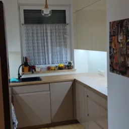 Jasna kuchnia z beżowymi frontami szafek, drewnianym blatem, czarnym zlewem i oknem z koronkową firanką, oświetlona wiszącą lampą i podświetleniem podszafkowym.