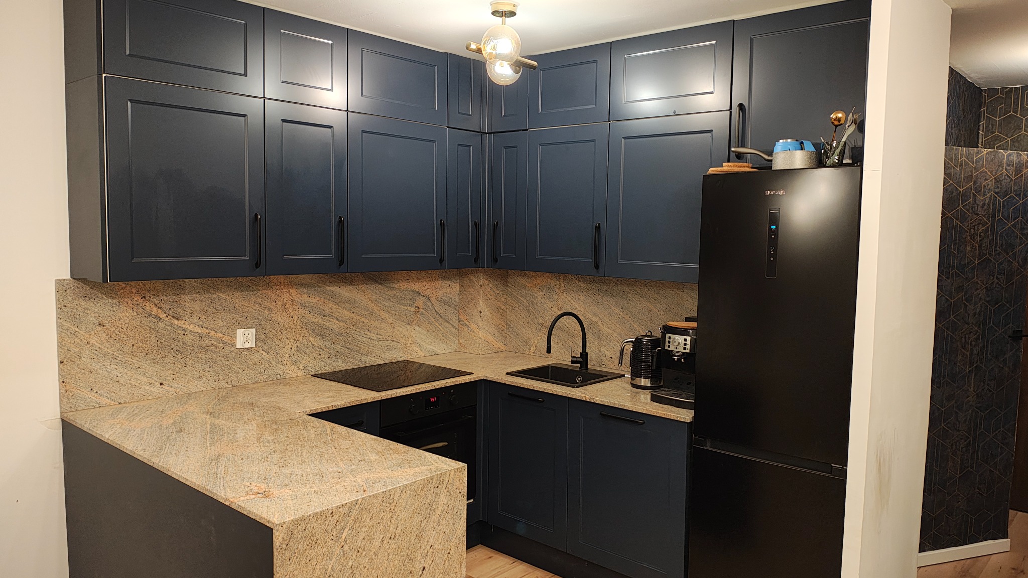 Granatowa kuchnia na wymiar z blatem z kamienia o ciepłej barwie, widoczna płyta indukcyjna, czarny zlew i bateria, podwieszane szafki z długimi uchwytami, nowoczesna lampa sufitowa.