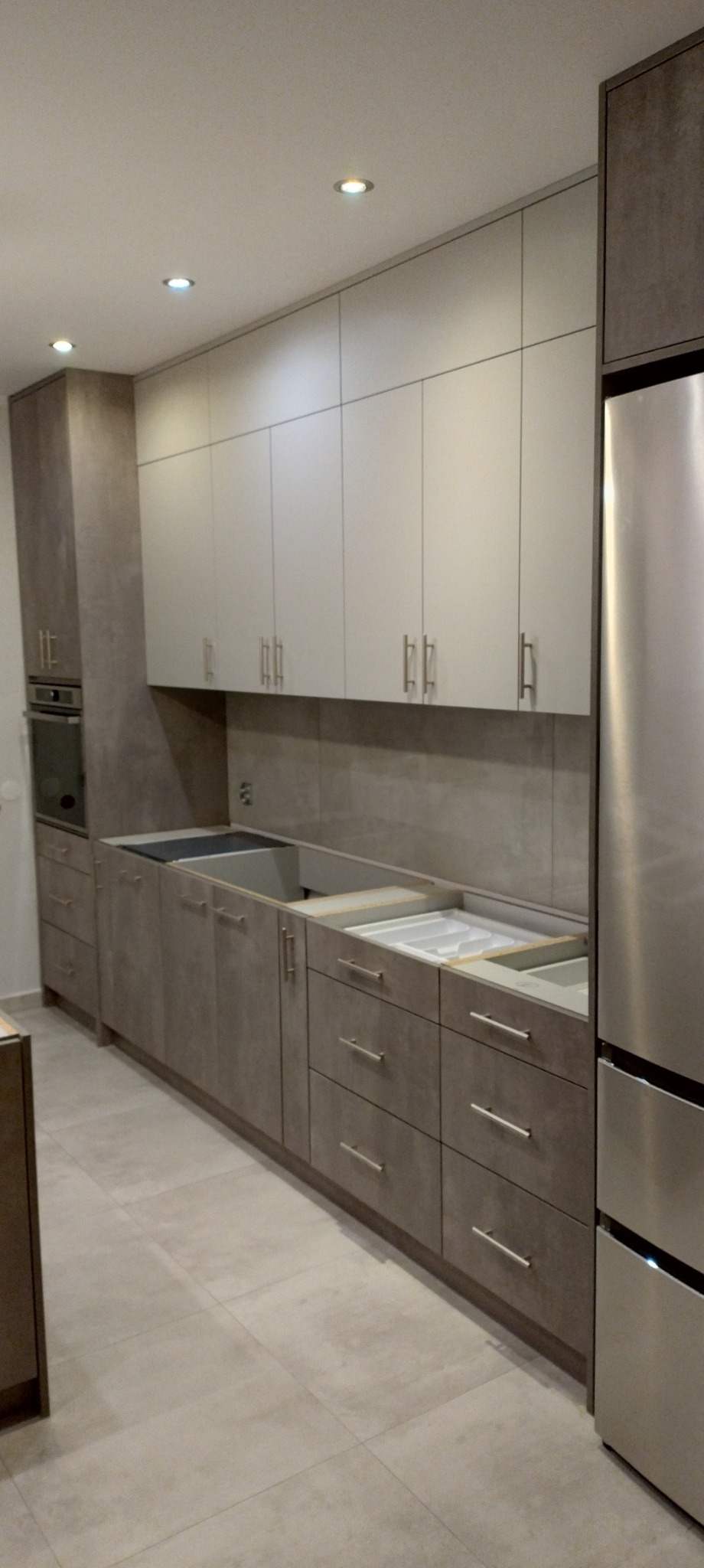 Wnętrze kuchni w trakcie montażu: szare dolne szafki z srebrnymi uchwytami, białe górne szafki, wstawione zlewozmywaki bez baterii, obok lodówka ze stali nierdzewnej.