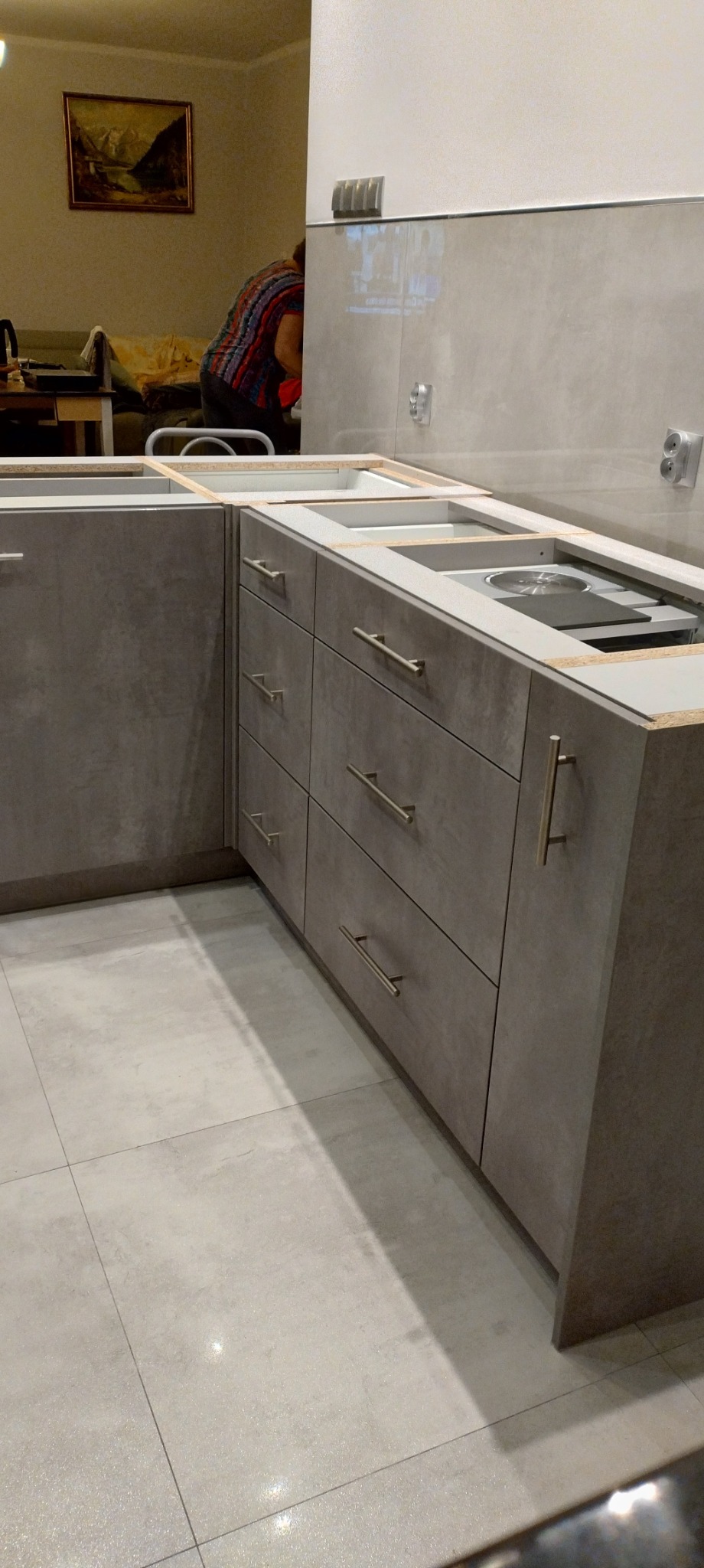 Montaż szarej kuchni z szufladami i miejscem na płytę grzewczą, widoczne niedokończone blaty i fragment pomieszczenia z obrazem na ścianie.