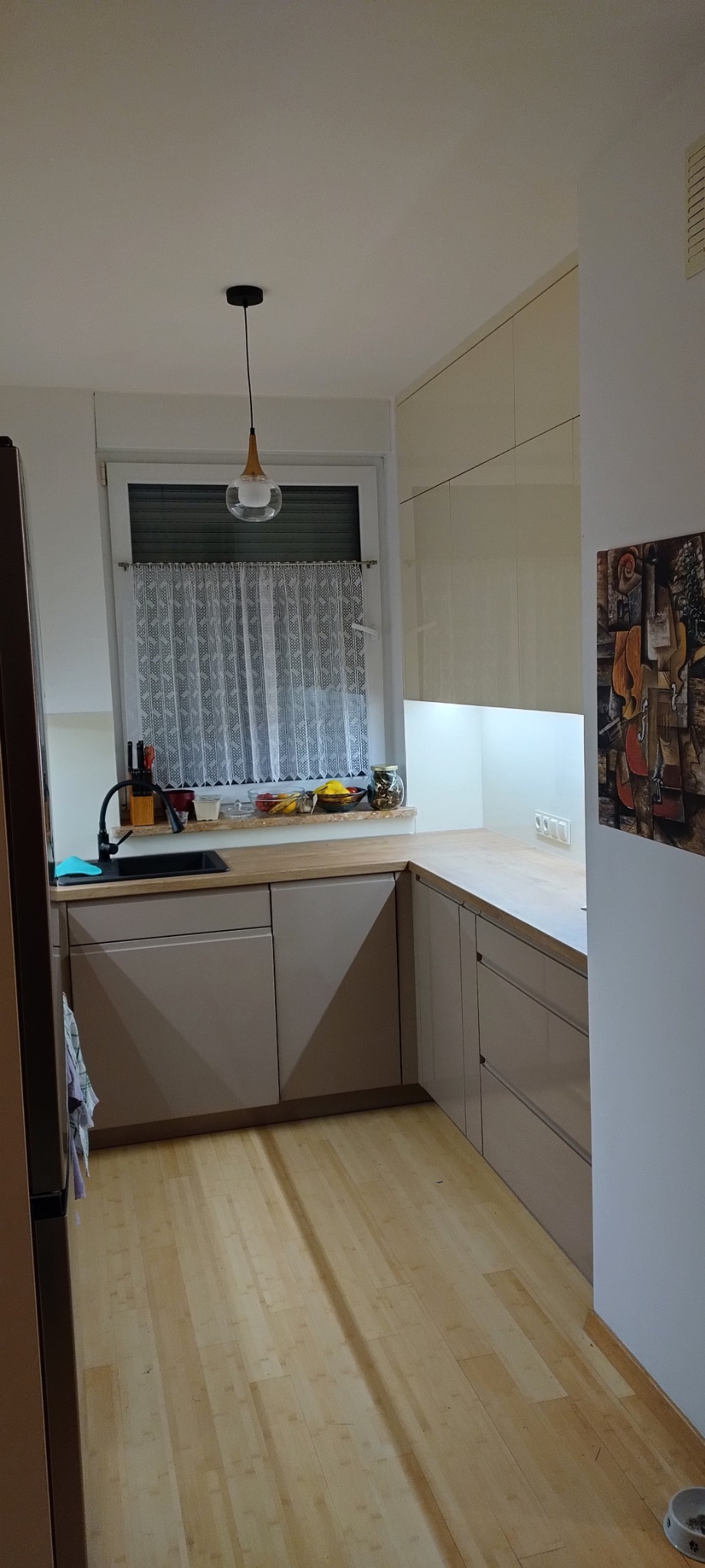 Jasna kuchnia z beżowymi frontami szafek, drewnianym blatem, czarnym zlewem i oknem z koronkową firanką, oświetlona wiszącą lampą i podświetleniem podszafkowym.