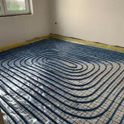 Niebieskie rury systemu ogrzewania podłogowego ułożone spiralnie na srebrnej folii izolacyjnej z nadrukowaną siatką, widoczne żółte taśmy przy ścianach i fragment okna.