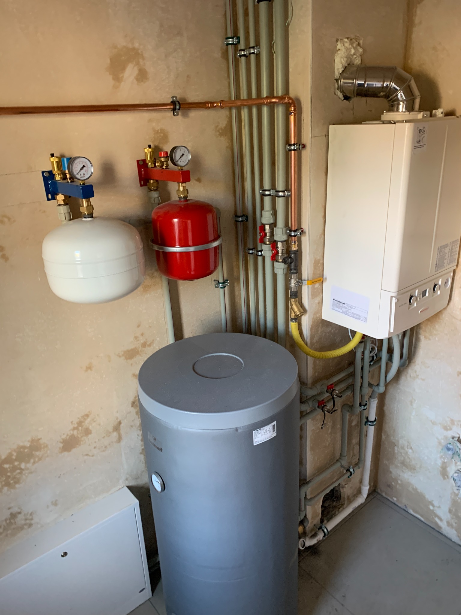 Kocioł grzewczy wiszący z podłączeniami, zasobnik ciepłej wody, naczynia przeponowe w instalacji centralnego ogrzewania, widoczne rury miedziane i plastikowe, manometry na zbiornikach, ściana...