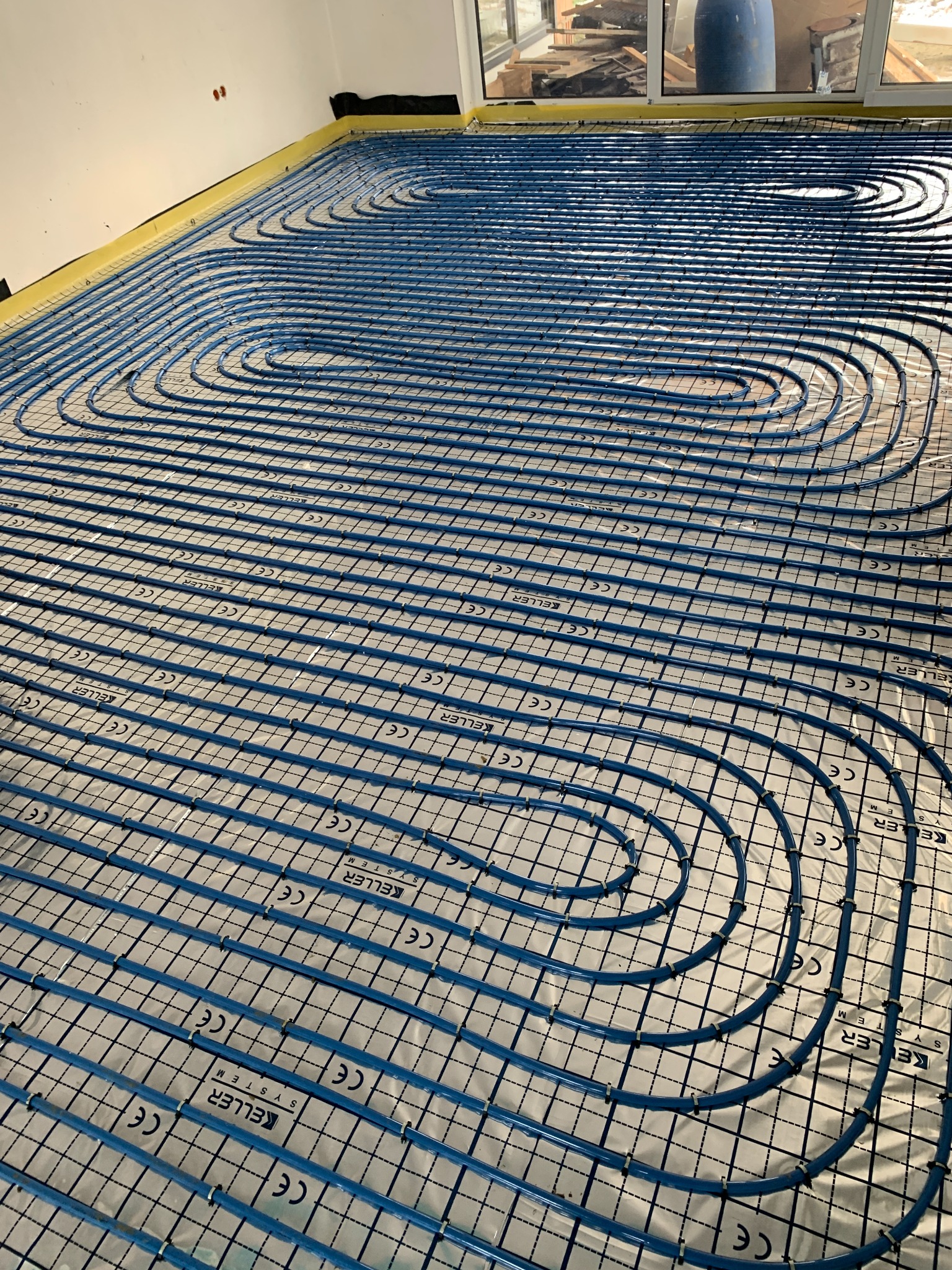 Niebieskie rury spiralnie ułożone na srebrnej folii z nadrukiem siatki i logo Keller System, przygotowane do zalania wylewką ogrzewania podłogowego, widoczne elementy izolacji przy ścianach.