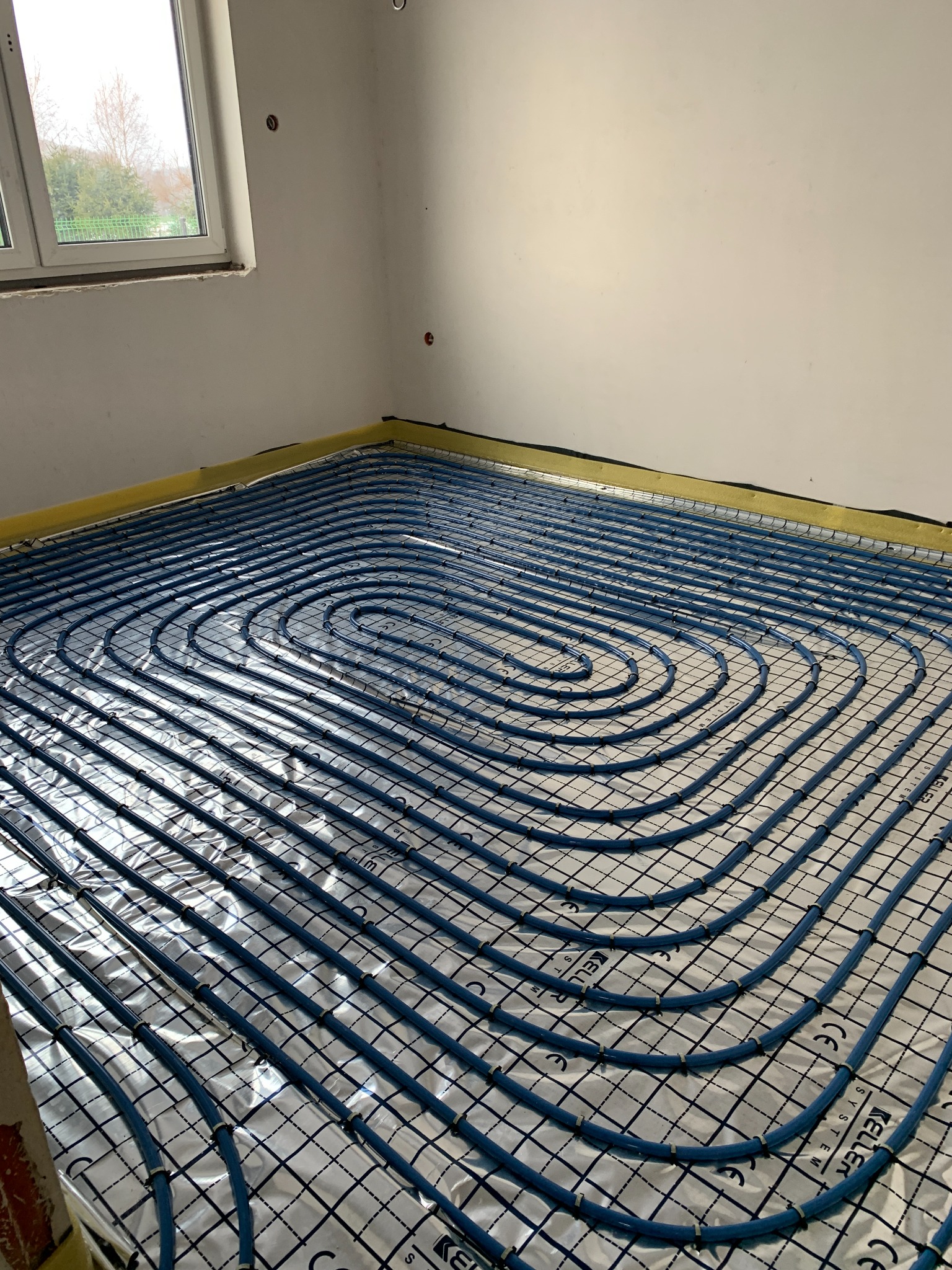 Niebieskie rury systemu ogrzewania podłogowego ułożone spiralnie na srebrnej folii izolacyjnej z nadrukowaną siatką, widoczne żółte taśmy przy ścianach i fragment okna.