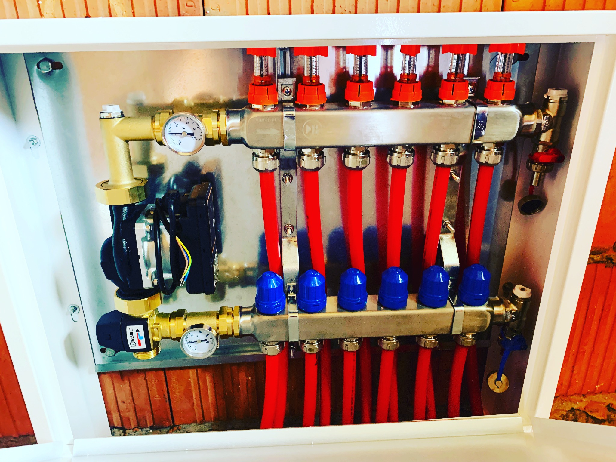 Rozdzielacz ogrzewania podłogowego w szafce, widoczne czerwone rury, zawory z niebieskimi i pomarańczowymi pokrętłami, manometry oraz pompa obiegowa, tło z cegły.