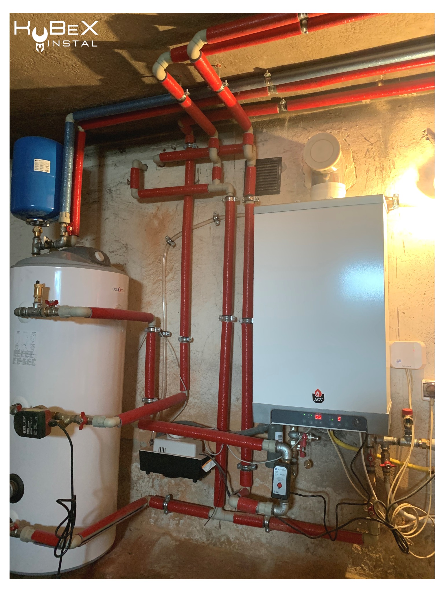 Kotłownia z piecem grzewczym ACV, bojlerem Golotto i rozbudowaną instalacją z rur w izolacji termicznej, widoczne liczne zawory i czujniki, całość w piwnicy o surowych ścianach.