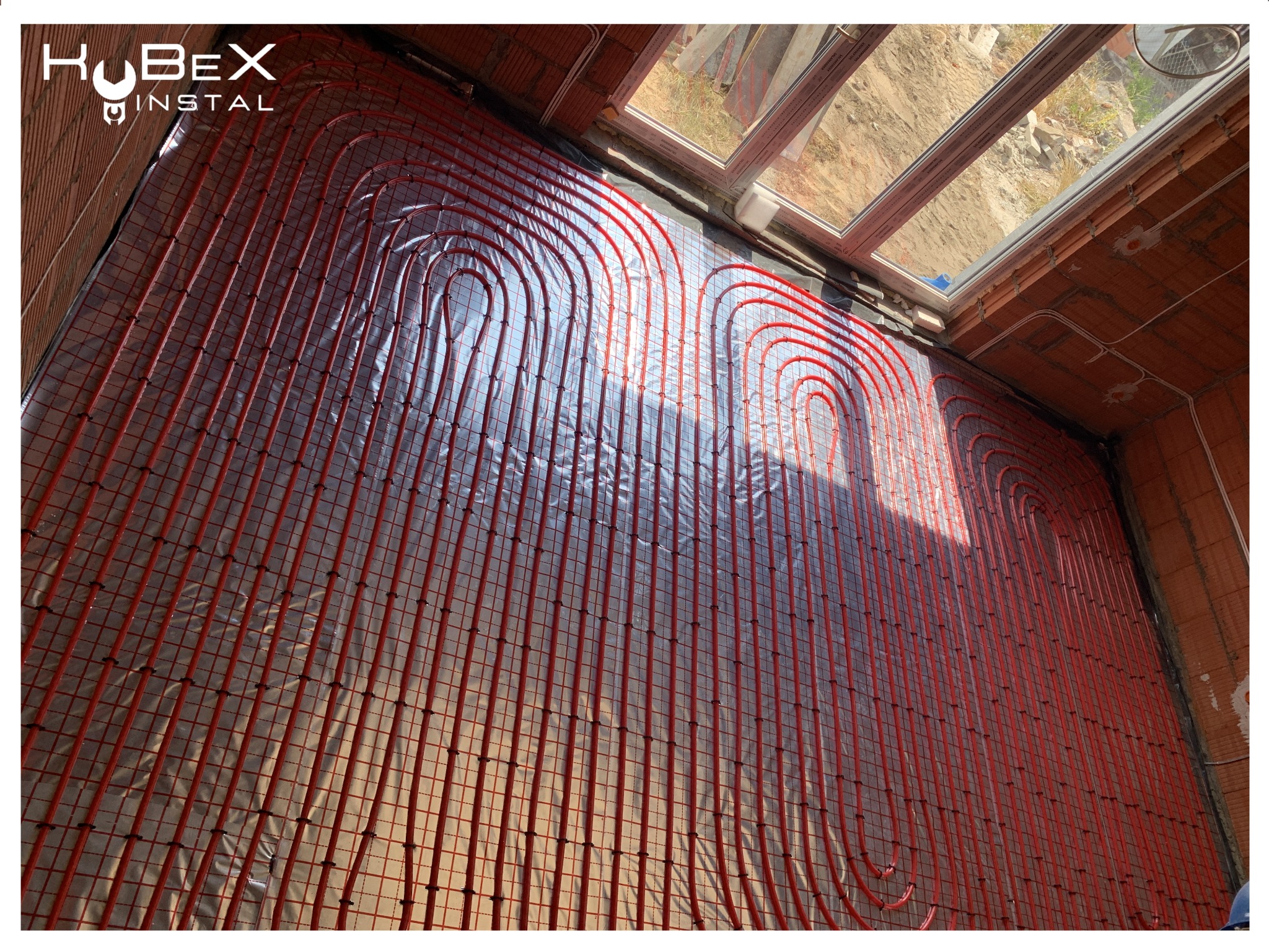 Czerwone rurki ogrzewania podłogowego ułożone na srebrnej folii izolacyjnej w surowym wnętrzu z ceglanymi ścianami i oknem dachowym, widoczne logo firmy Kubex Instal.