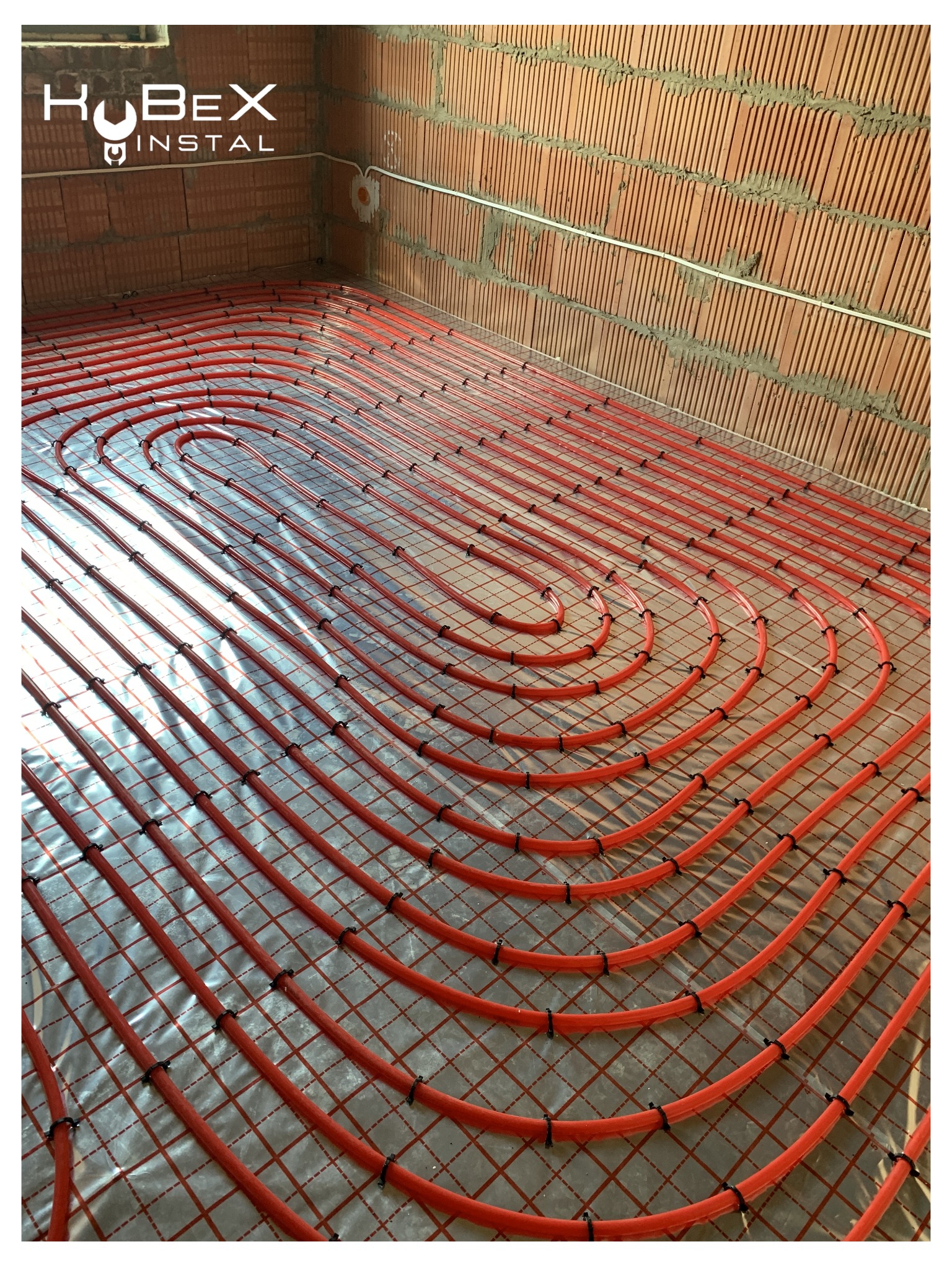 Czerwone rury równolegle ułożone na srebrnej folii z siatką w surowym wnętrzu z ceglanymi ścianami, widoczne logo firmy Kubex Instal.
