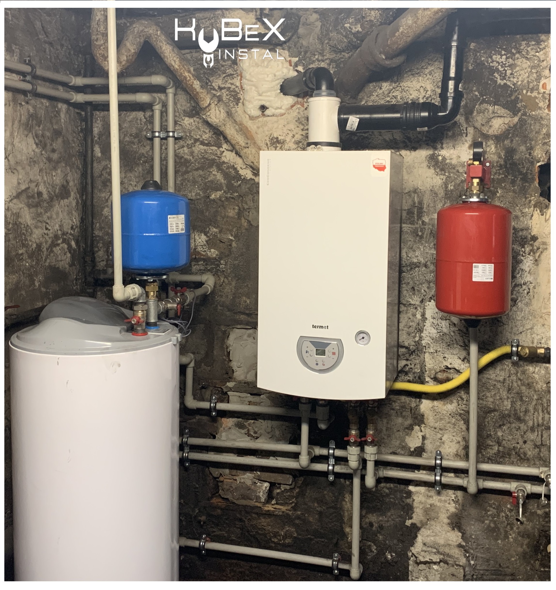 Kotłownia z piecem gazowym Termet, zbiornikiem CWU z niebieskim naczyniem przeponowym, czerwonym naczyniem przeponowym i instalacją hydrauliczną na tle surowej, kamiennej ściany piwnicy. Widoczne...