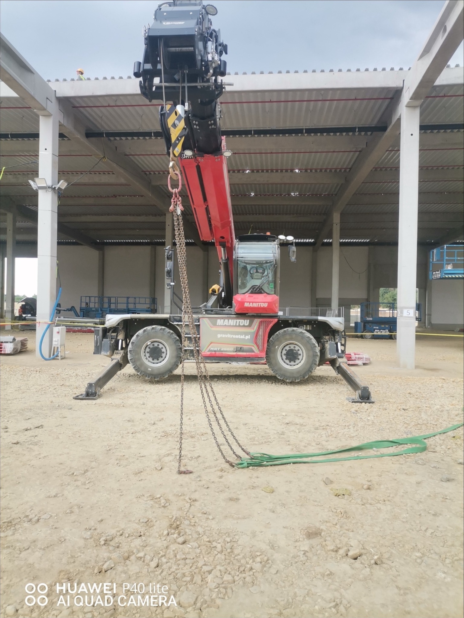 Czerwony dźwig samojezdny Manitou na placu budowy hali stalowej, widoczny pracownik na dachu, w tle podnośniki koszowe.