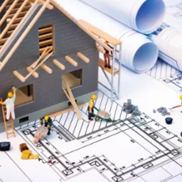 Miniaturowy model domu w trakcie budowy, z figurkami robotników, betoniarką i taczką, umieszczony na planie architektonicznym, z rolkami rysunków w tle.