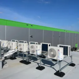 Rząd jednostek zewnętrznych klimatyzacji marki Gree, zamontowanych na dachu hali przemysłowej, widoczne kable i konstrukcja wsporcza.