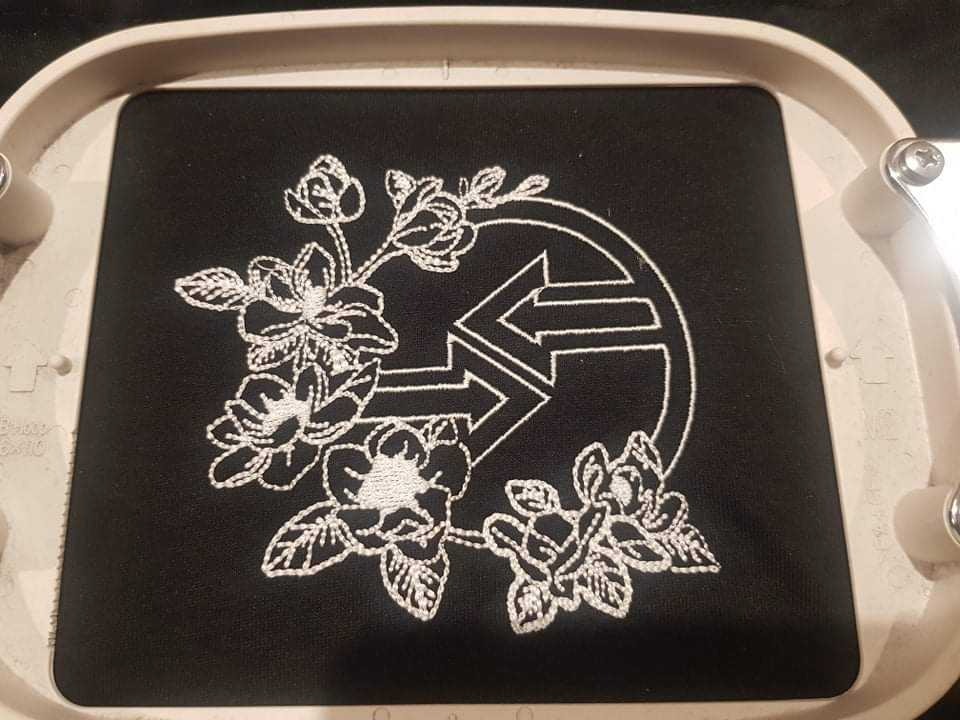 Czarna tkanina w tamborku z białym haftem przedstawiającym stylizowane kwiaty i logo z podwójną strzałką.