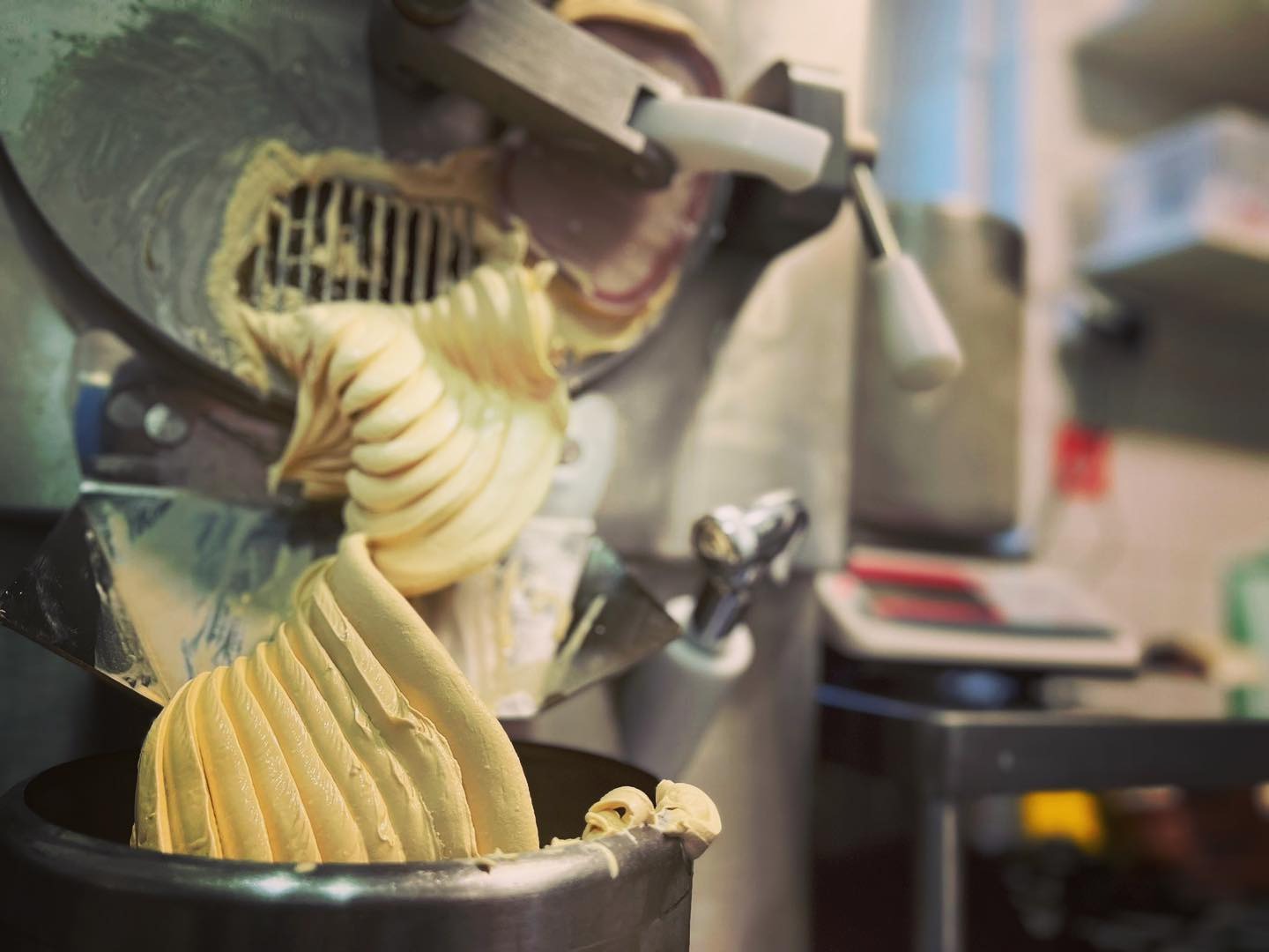 Automat do lodów w trakcie pracy, kremowy deser wyciskany do metalowego pojemnika, w tle widoczne elementy wyposażenia kuchennego.