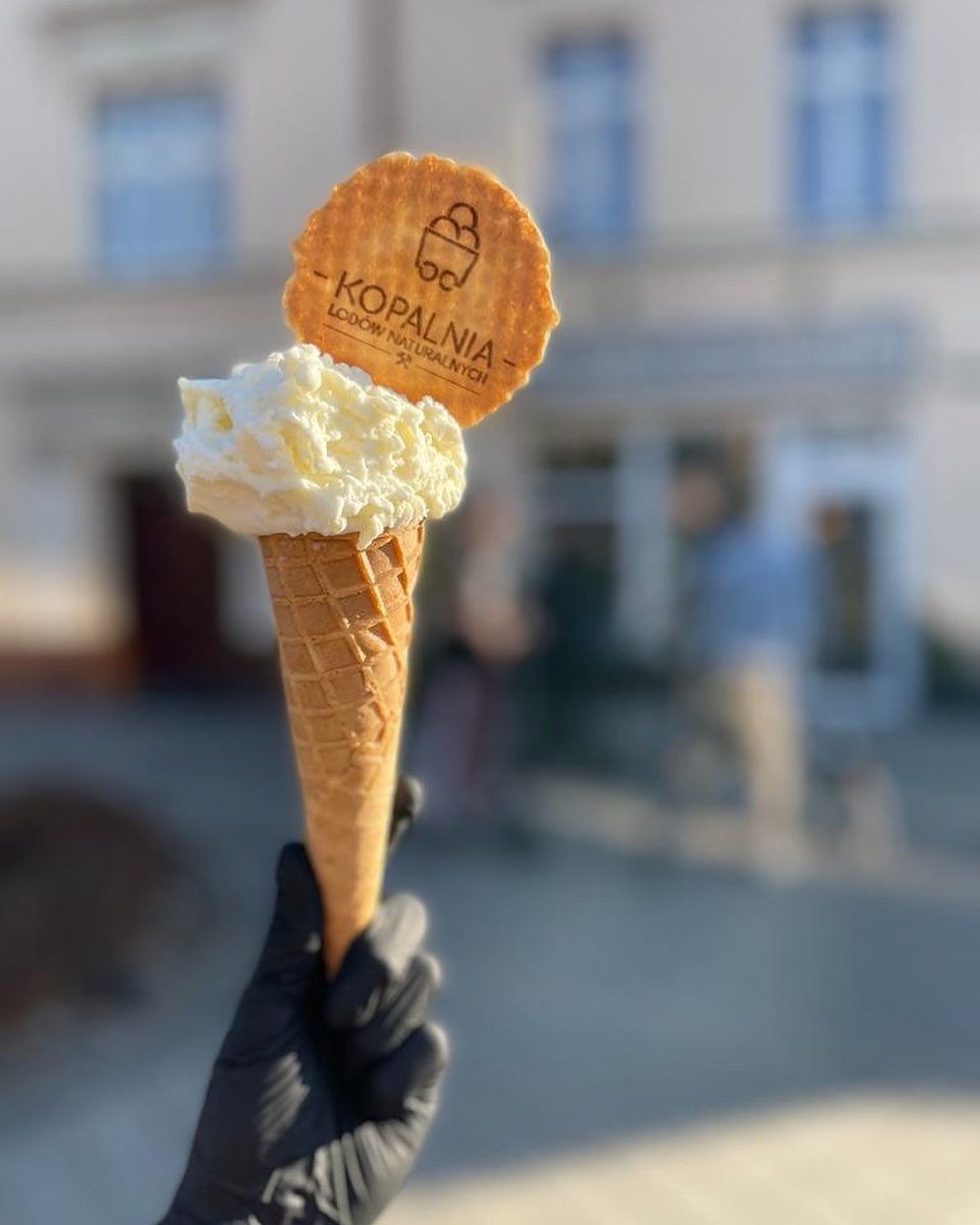 Wafel z logo na gałce lodów waniliowych w wafelku, trzymany w dłoni w czarnej rękawiczce, na rozmytym tle budynku.