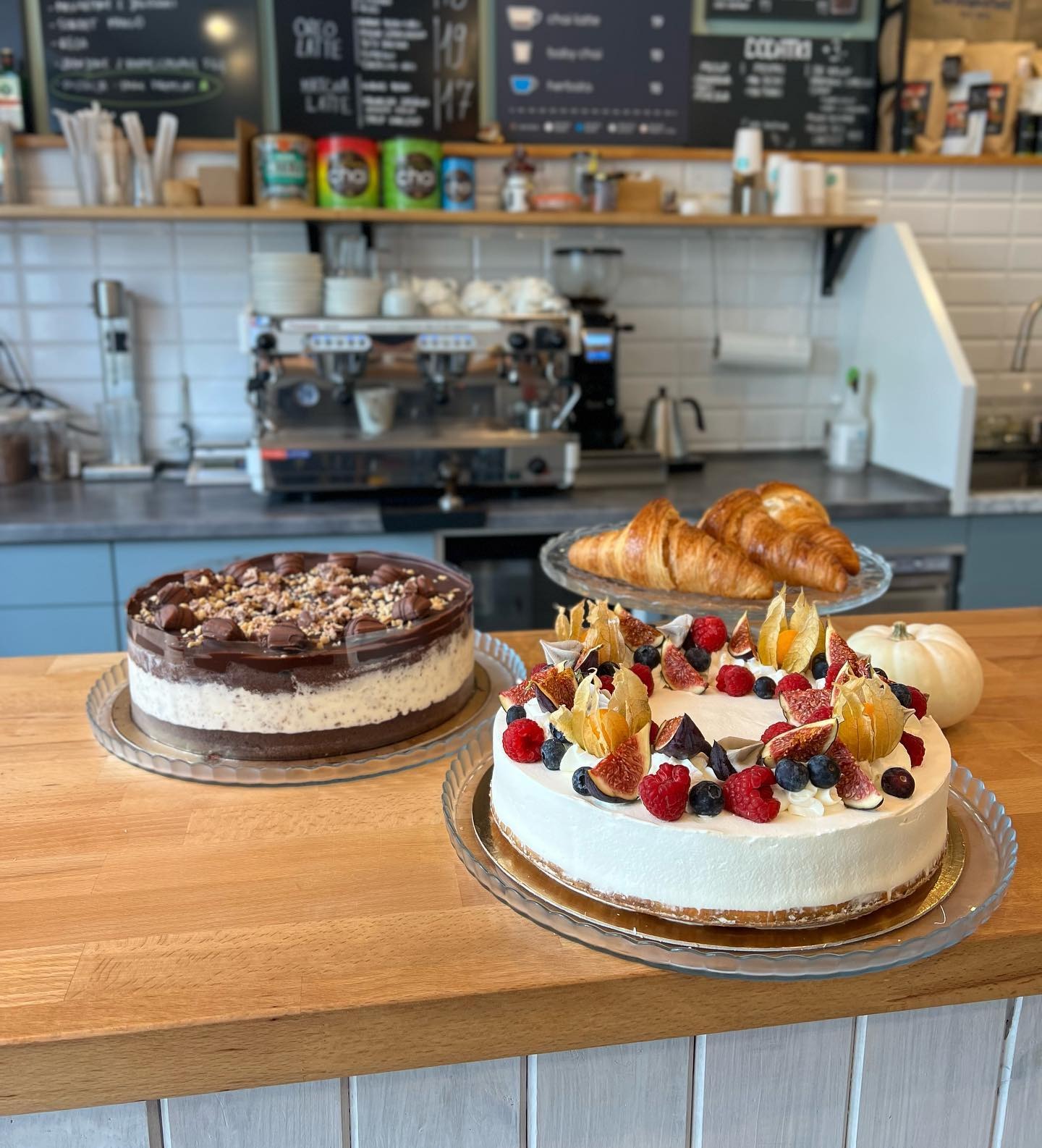 Dwa ciasta na drewnianym blacie, jedno z czekoladą i orzechami, drugie z kremem, figami, malinami i borówkami, w tle ekspres do kawy i croissanty.