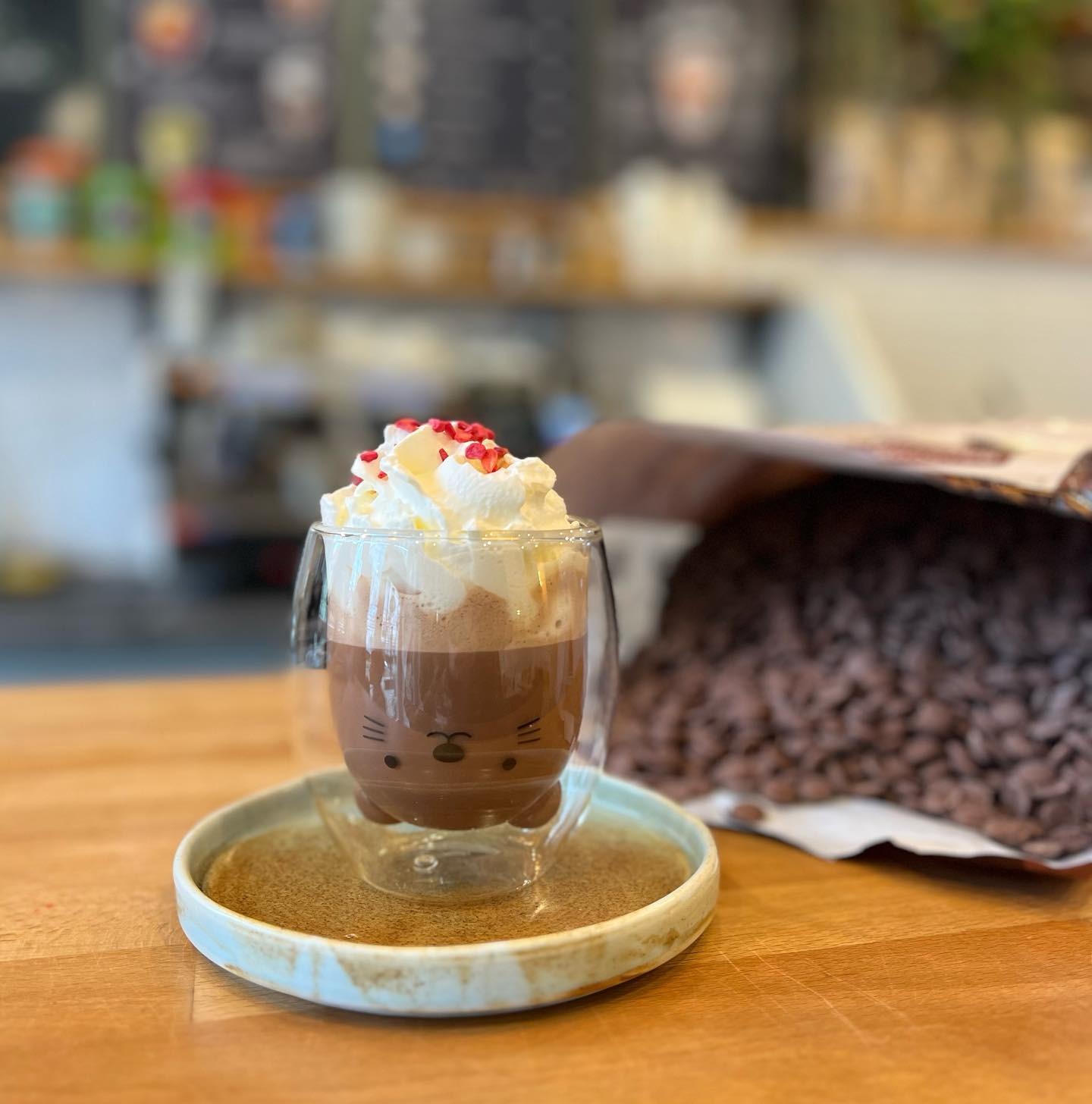 Gorąca czekolada w szklance z motywem zwierzęcym, udekorowana bitą śmietaną i posypką z czerwonych owoców, na ceramicznej podstawce, obok otwartego worka z czekoladowymi kroplami.