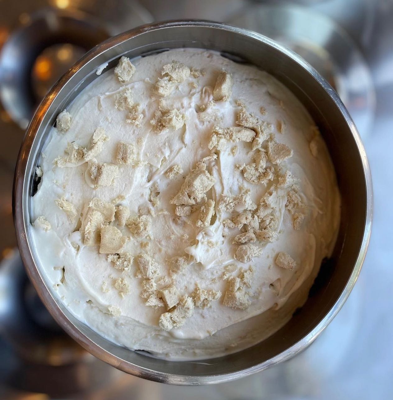 Widok z góry na metalową misę wypełnioną kremem o białej barwie, posypanym nieregularnymi kawałkami beżowej kruszonki, prawdopodobnie deser lub składnik ciasta w trakcie przygotowywania.