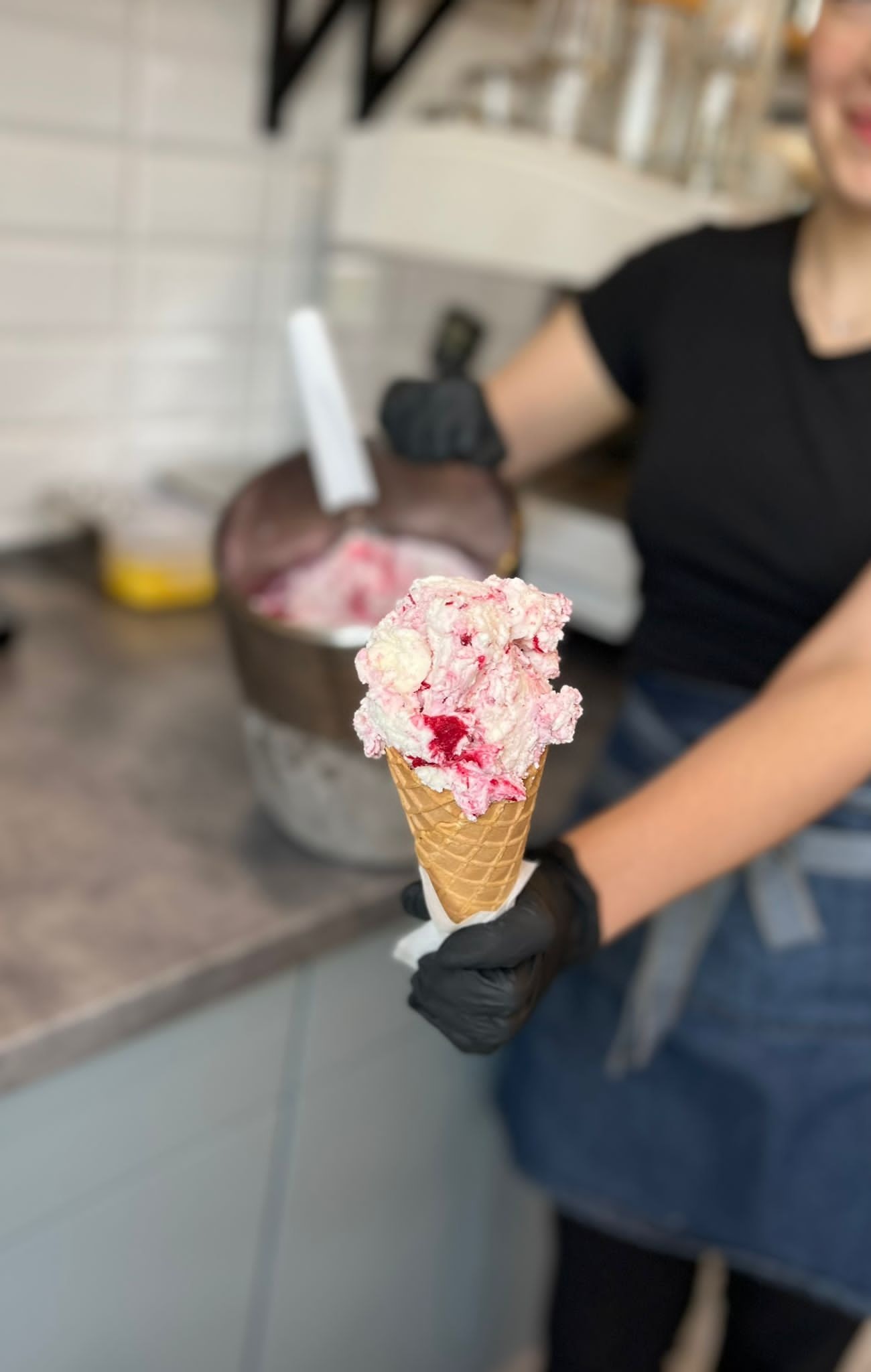 Kremowe lody truskawkowe w wafelku, podawane przez osobę w czarnych rękawiczkach i dżinsowym fartuchu, z metalowym pojemnikiem z lodami w tle.