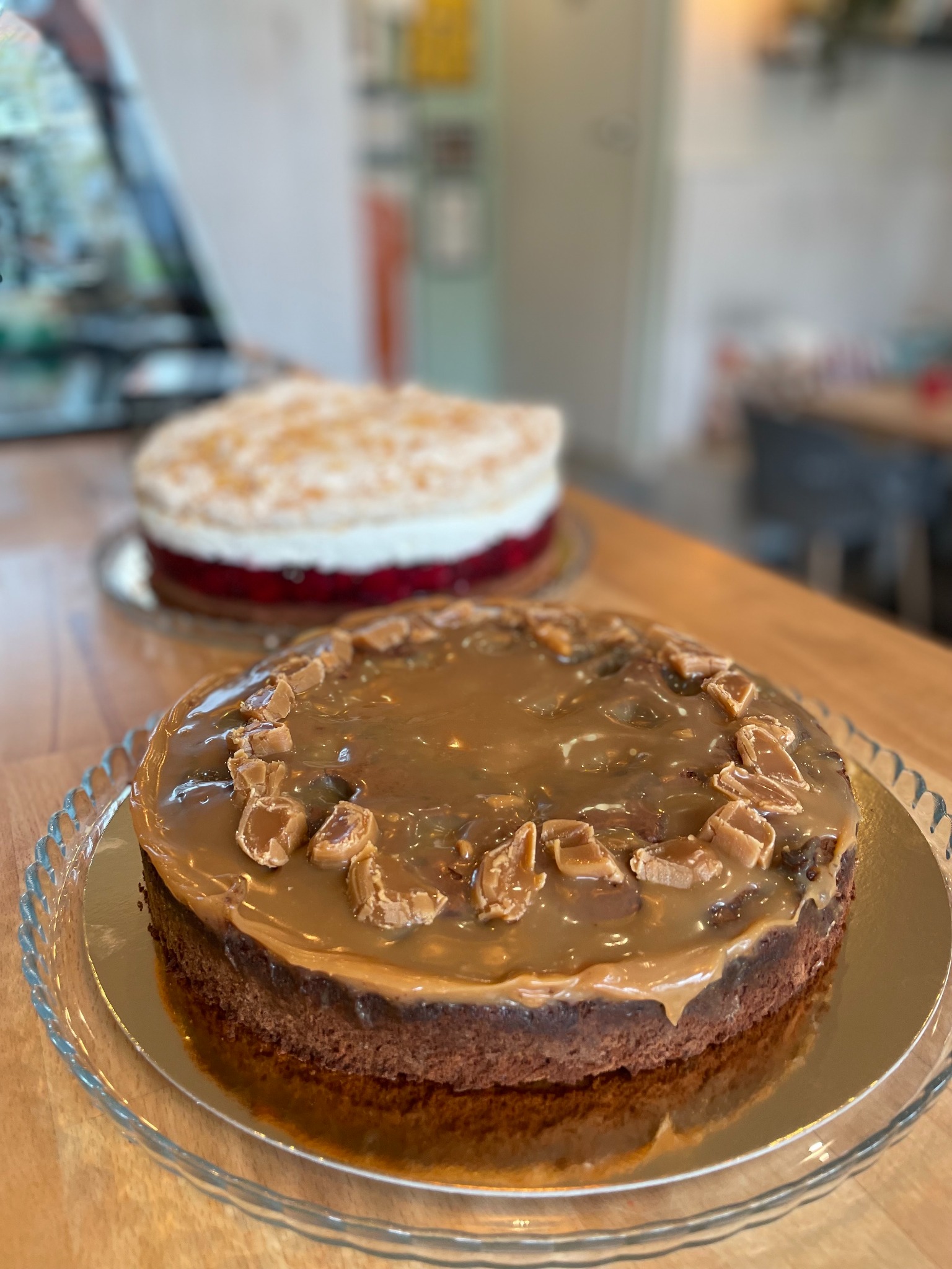 Ciemny tort czekoladowy polany karmelem i udekorowany kawałkami karmelków, na szklanym talerzu na nóżce, w tle drugi tort z bitą śmietaną i galaretką.