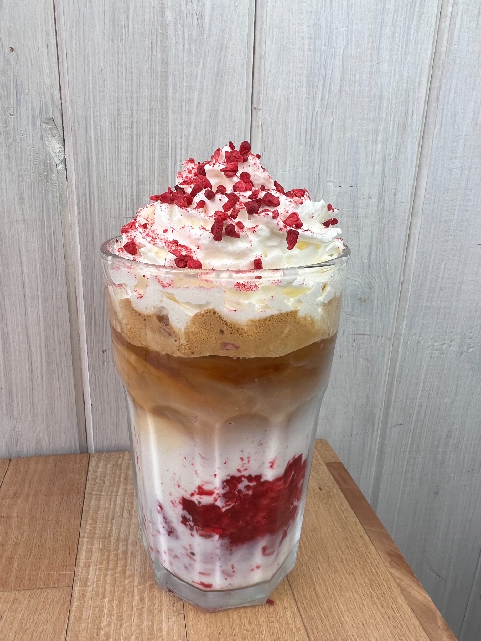 Wysoka szklanka z warstwowym deserem: na dnie czerwone owoce zatopione w mleku, następnie warstwa kawy, całość zwieńczona bitą śmietaną posypaną kawałkami czerwonych owoców. Deser stoi...