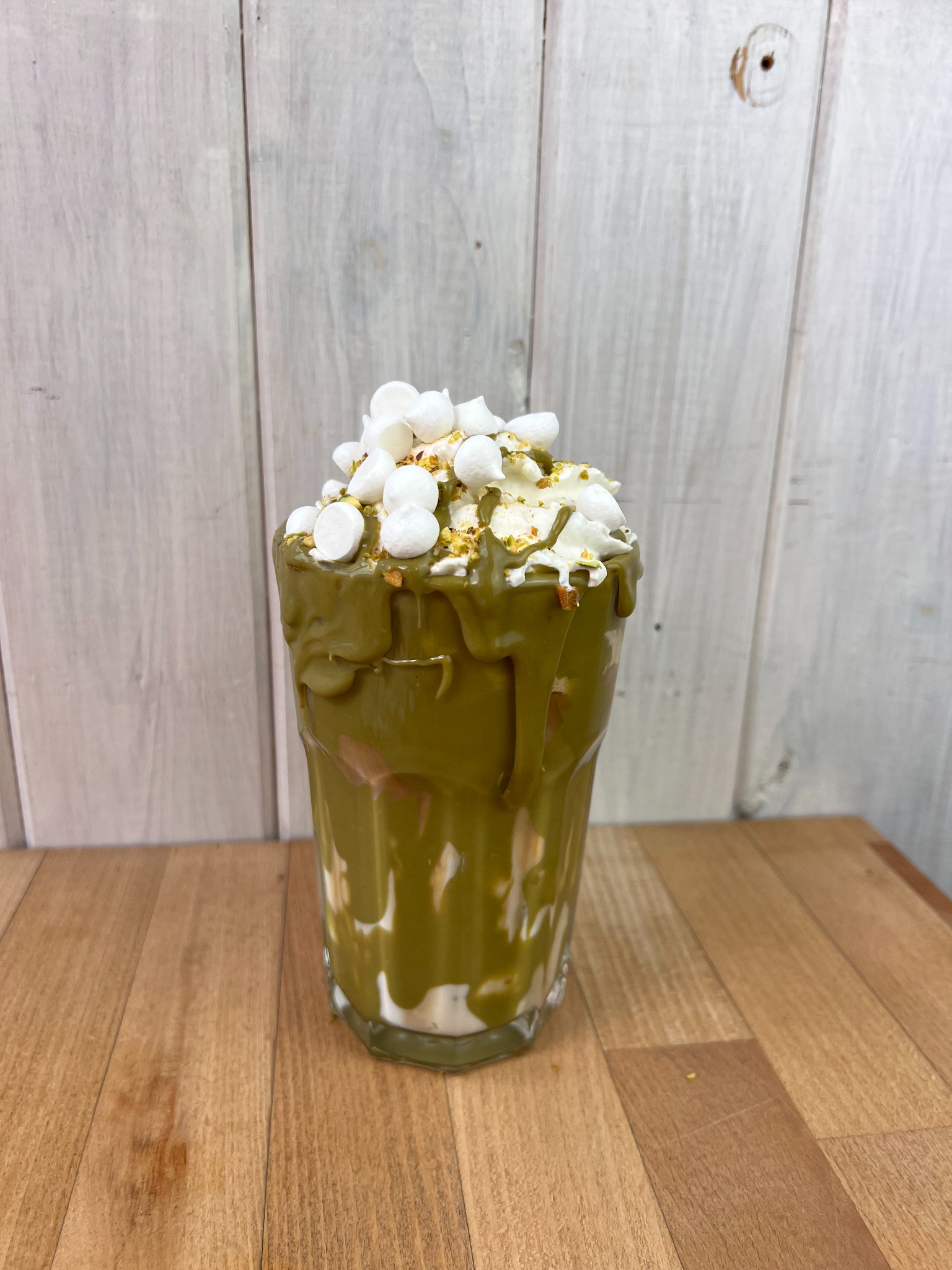 Deser w szklance z zieloną polewą, bitą śmietaną i mini piankami marshmallow, posypany pistacjami na drewnianym stole i tle z desek.