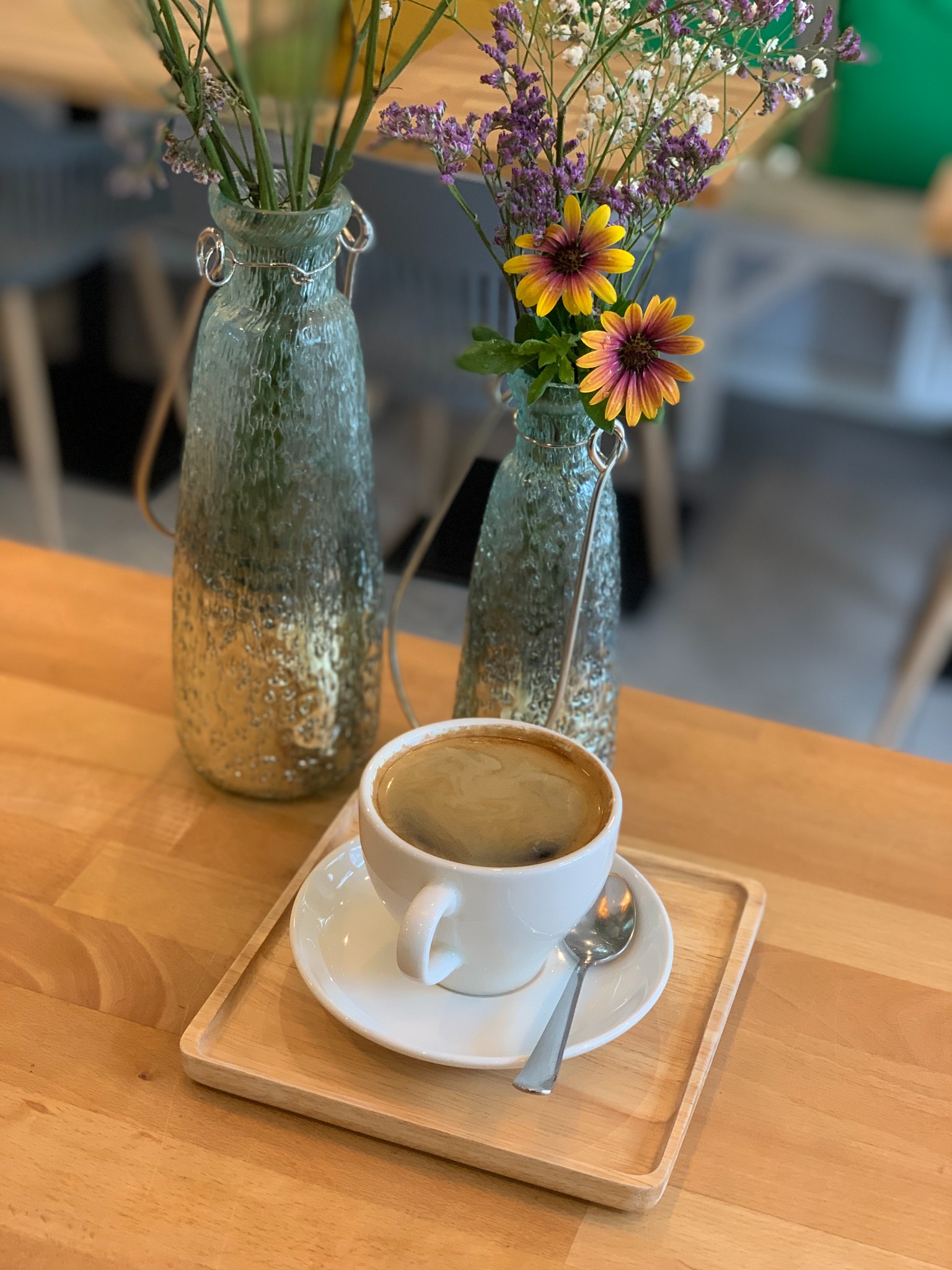 Filiżanka kawy na drewnianej tacy, obok dwie szklane butelki z kwiatami na drewnianym stole.