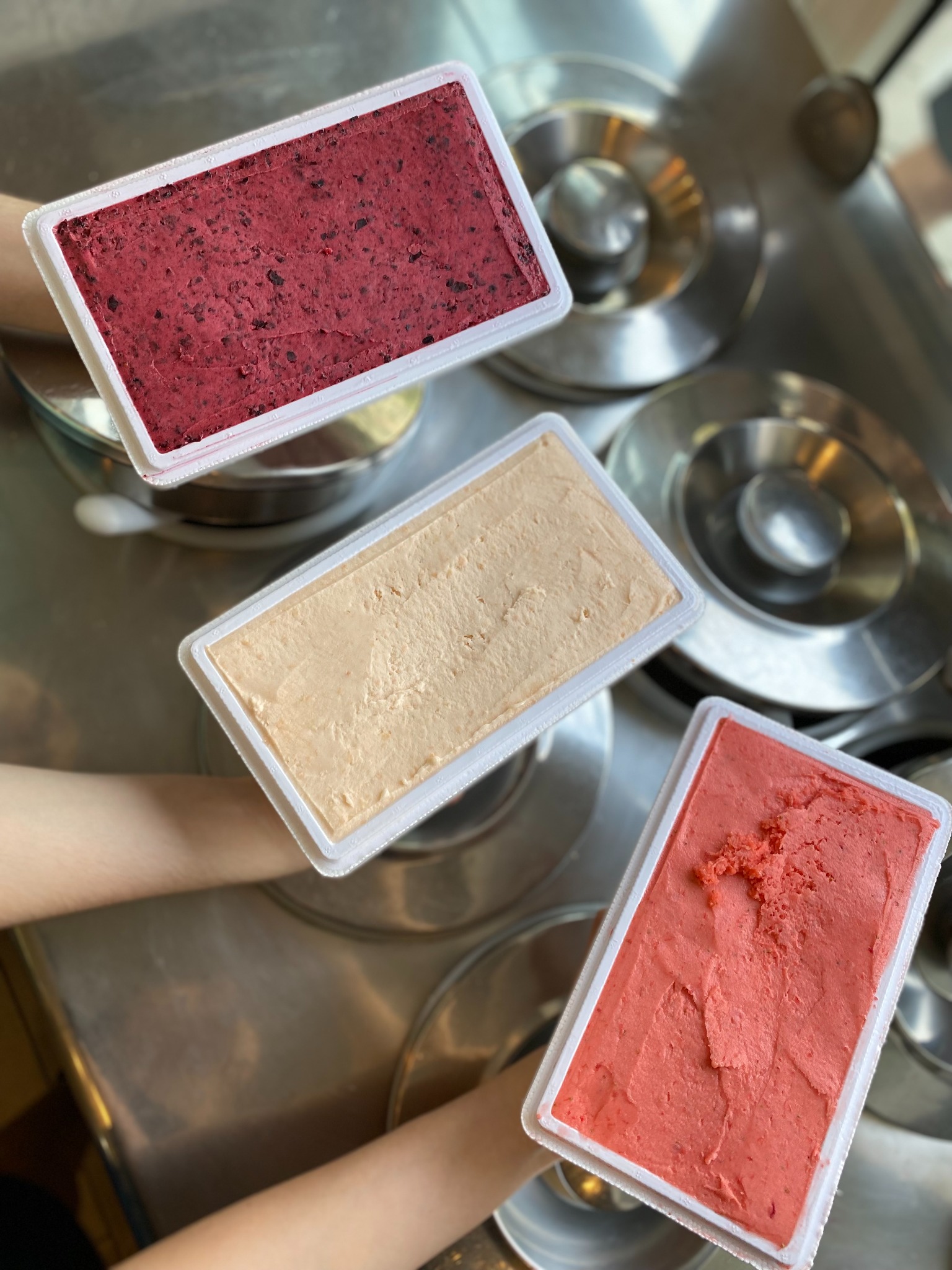 Trzy prostokątne pojemniki z lodami o różnych smakach (jagodowe, brzoskwiniowe i truskawkowe) trzymane nad metalową powierzchnią z okrągłymi wgłębieniami.