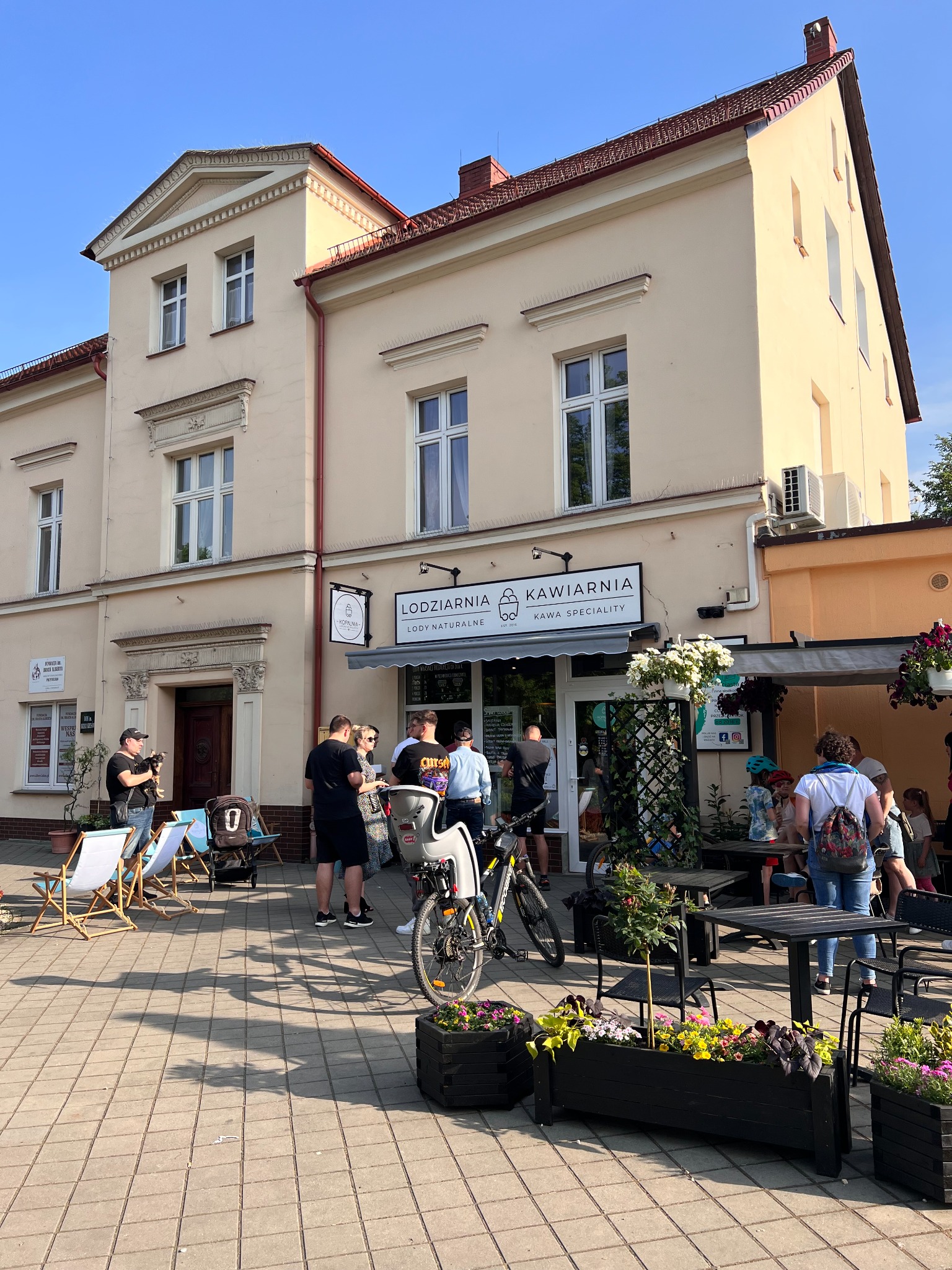 Elewacja budynku z szyldem 'Lodzarnia Kawiarnia' nad wejściem, przed którym stoją klienci i stoliki z kwiatami, a obok leżaki.