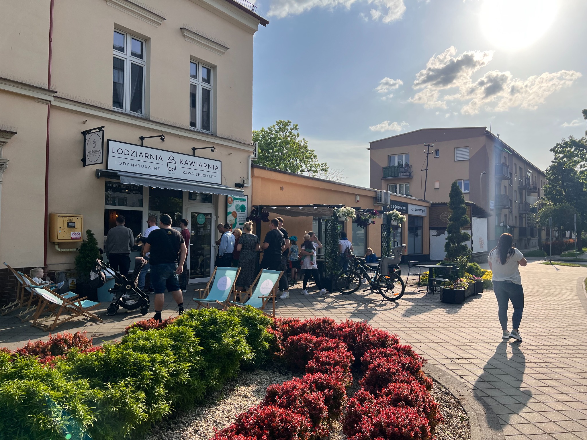 Kolejka klientów przed lodziarnią-kawiarnią w słoneczny dzień. Na pierwszym planie rabata z czerwonymi krzewami, za nią leżaki i wózki dziecięce. W tle budynek z szyldem 'Lodziarnia Kawiarnia'...