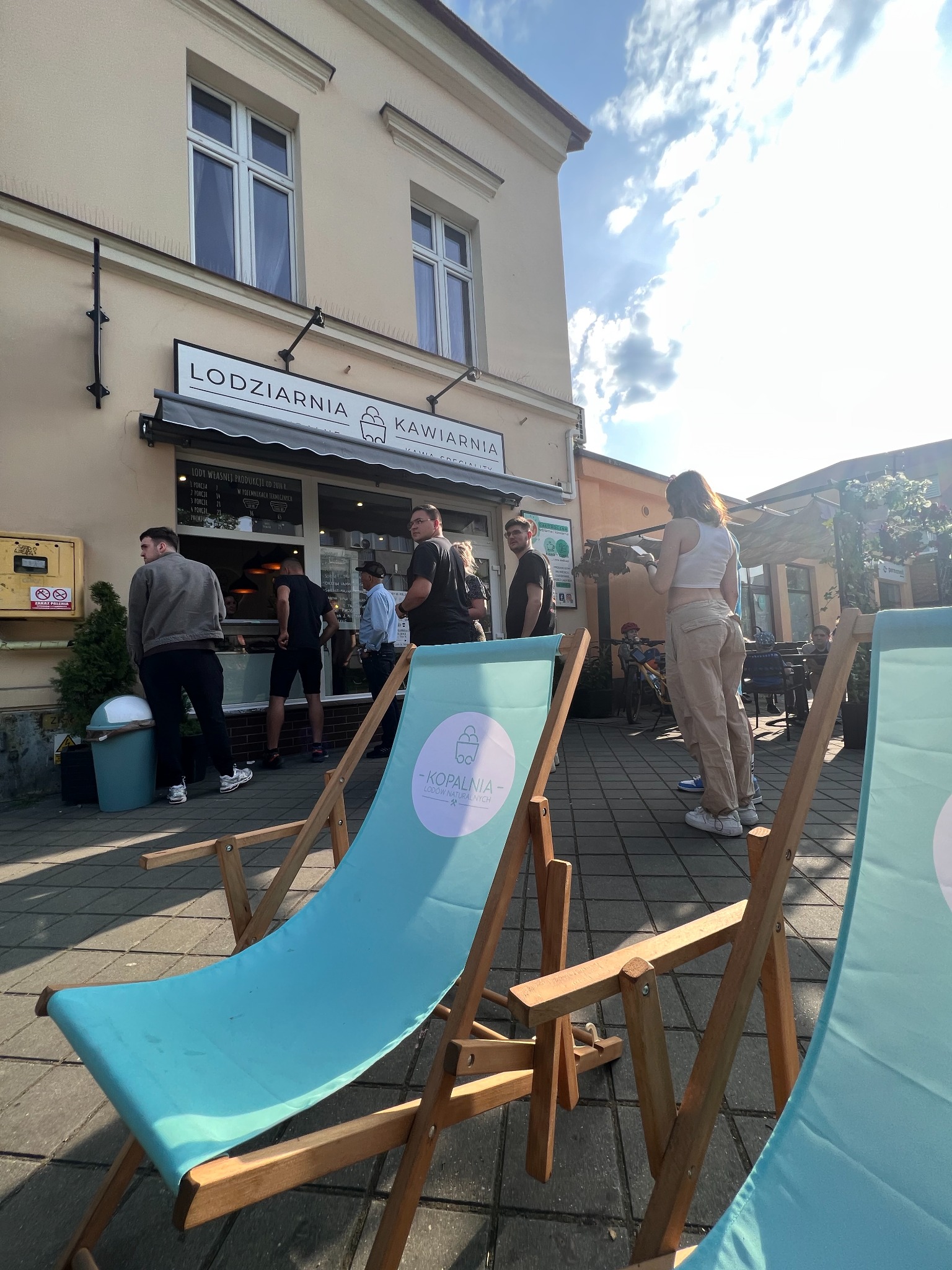 Kolejka klientów przed lodziarnią z niebieskimi leżakami na pierwszym planie, widoczny szyld 'Lodziarnia Kawiarnia' i menu lodów własnej produkcji.