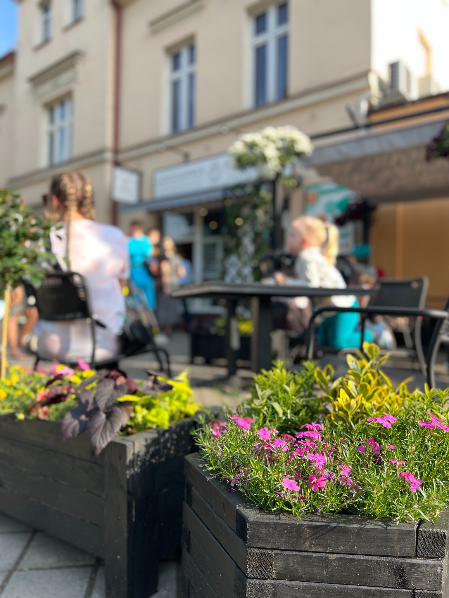 Kawiarniany ogródek z drewnianymi donicami pełnymi kolorowych kwiatów na pierwszym planie, w tle stoliki z klientami i fasada budynku.