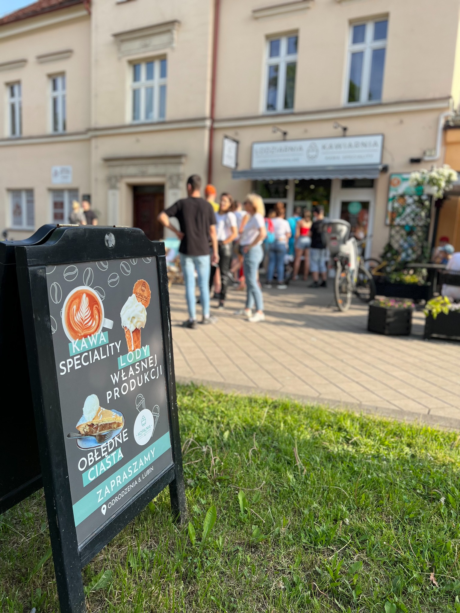 Czarny, dwustronny szyld reklamowy na trawniku przed lokalem z napojami i deserami; na szyldzie grafiki kawy, lodów i ciasta oraz napisy: Kawa speciality, Lody własnej produkcji, Obłędne ciasta...