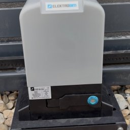 Smart Home Story - Szary napęd do bramy przesuwnej ELEKTROBIM KSE180-DG, zamontowany na czarnej podstawie wśród jasnych kamieni, obok ciemnej bramy we Wrocławiu.