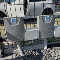 Smart Home Story - Dwa szare panele sterujące automatyki bramowej z logo ELEKTRO, zamontowane na czarnym słupku ogrodzenia, z kablami wychodzącymi z ziemi pokrytej żwirem.