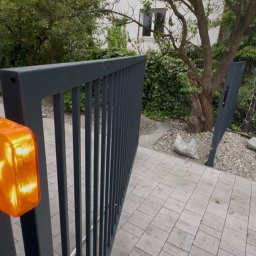 Smart Home Story - Częściowo otwarta szara brama przesuwna z pionowymi szczeblami i intensywnie świecącą pomarańczową lampą sygnalizacyjną na słupku, na tle zielonego ogrodu i brukowanego podjazdu.