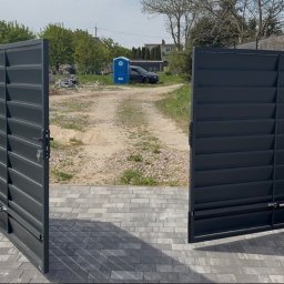 Smart Home Story - Otwarta, nowoczesna brama dwuskrzydłowa z ciemnoszarych lameli, z widocznymi siłownikami, na podjeździe z szarej kostki brukowej. W tle droga gruntowa i budowa.