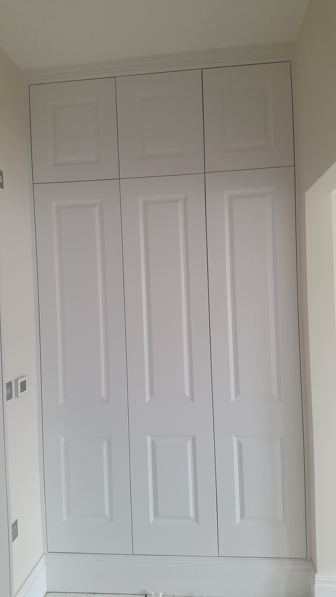 Biała, zabudowana szafa z sześcioma drzwiami w stylu klasycznym, z dekoracyjnymi panelami i listwami przypodłogowymi, dopasowana do wysokości pomieszczenia.
