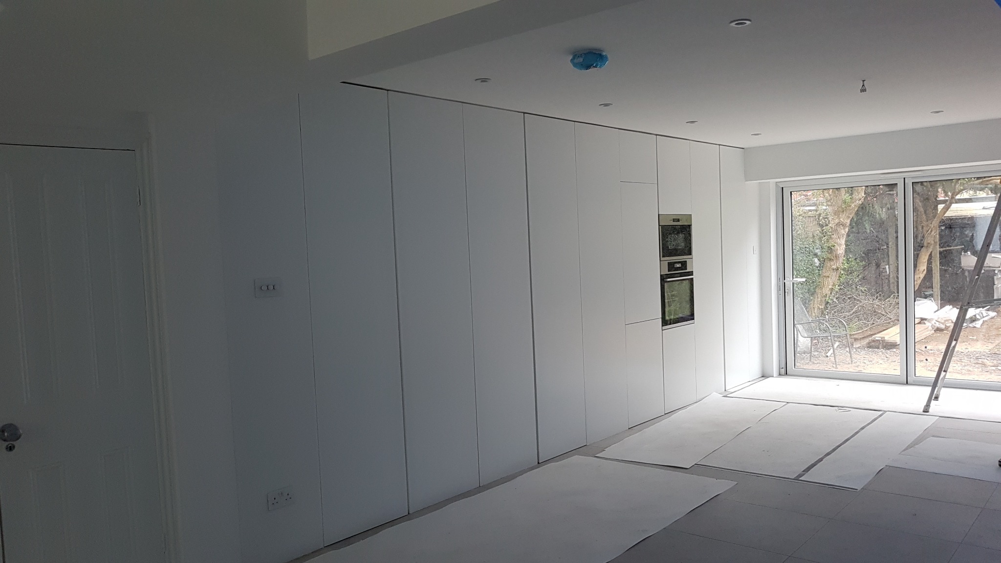 Wnętrze w trakcie wykańczania z zabudową meblową w kolorze białym, w której zamontowano piekarnik i mikrofalówkę. Na podłodze leżą kartony zabezpieczające. Widoczne drzwi balkonowe z widokiem...