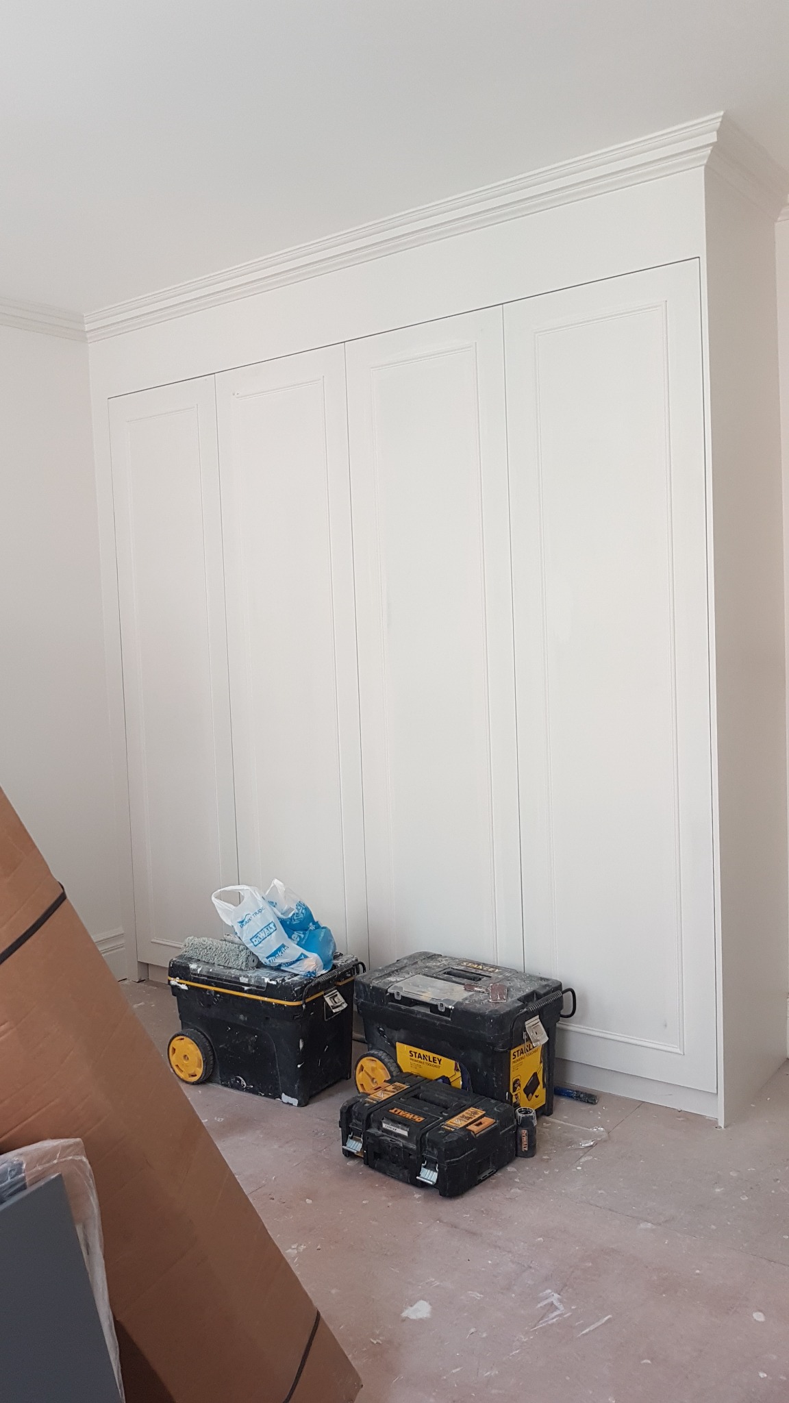 Biała szafa wnękowa z frezowanymi drzwiami, widoczne skrzynki narzędziowe Stanley i DeWalt na podłodze w trakcie montażu, zabezpieczone kartony w rogu pomieszczenia.