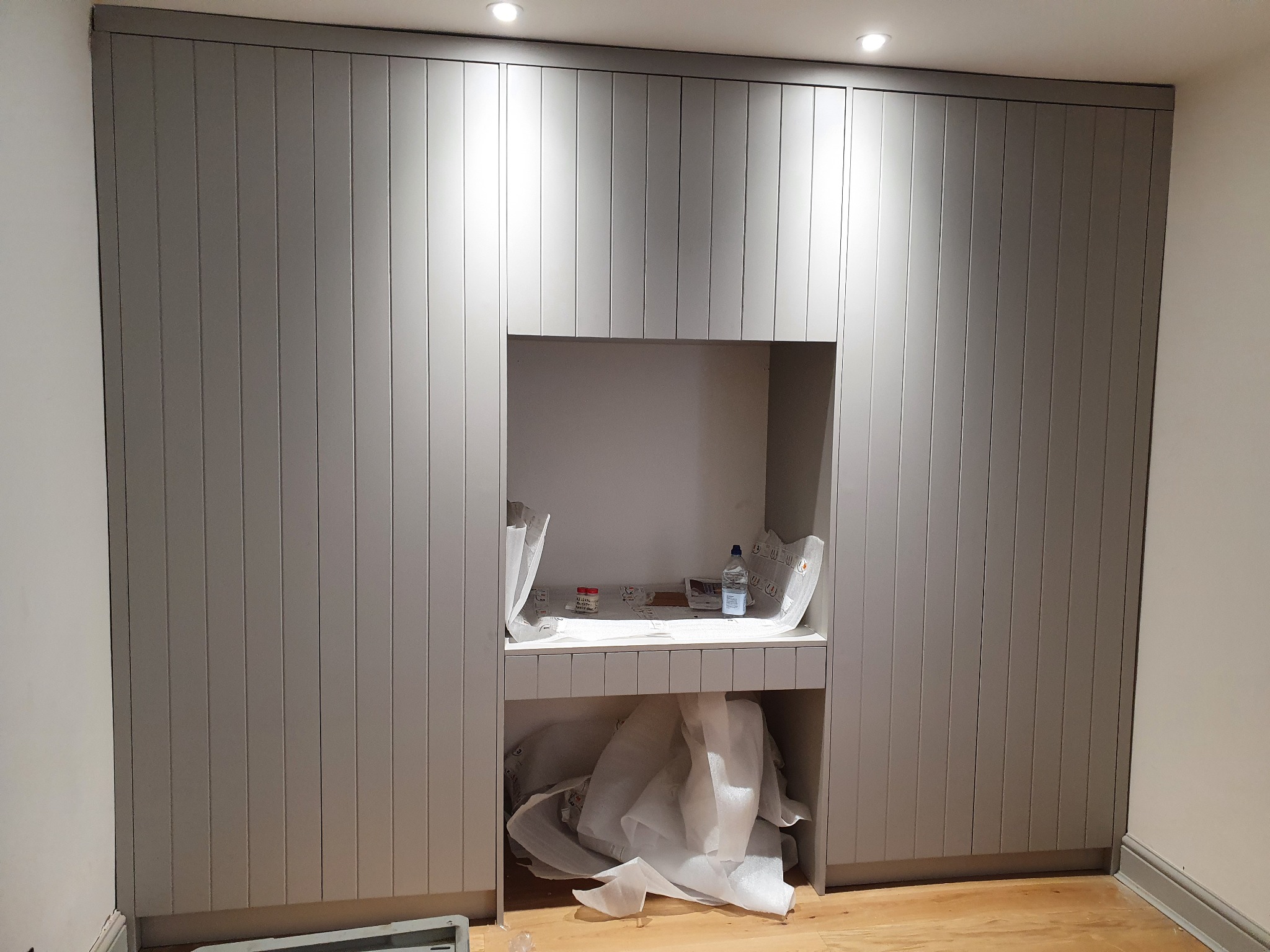 Szara zabudowa meblowa z pionowymi panelami i otwartą wnęką, w której znajdują się opakowania i butelka wody, podłoga drewniana, widoczne ślady po montażu.