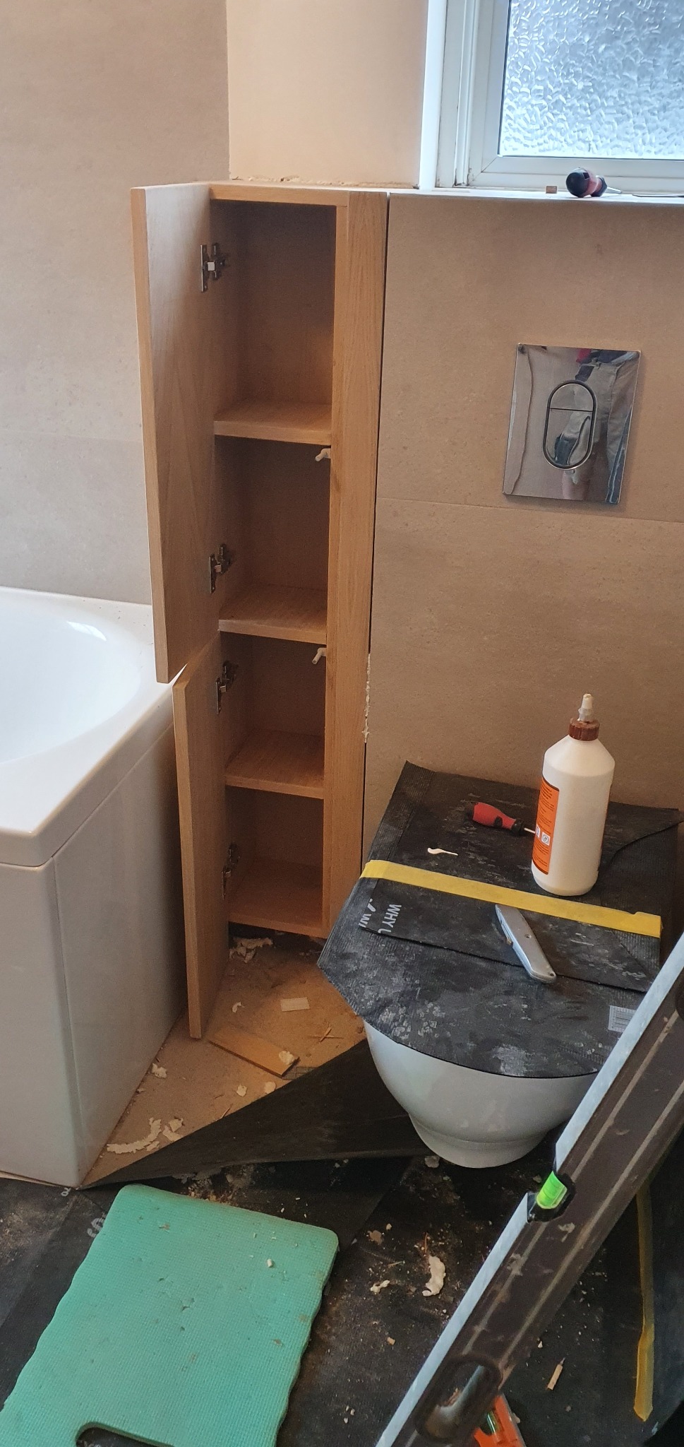 Wąska, drewniana szafka łazienkowa z otwartymi drzwiczkami, zamontowana obok wanny i przycisku spłuczki w trakcie instalacji, widoczne narzędzia, klej i resztki materiałów montażowych na podłodze.