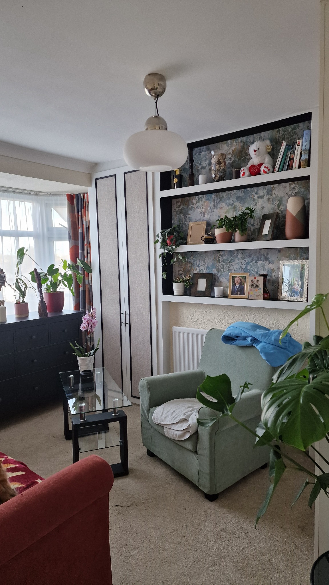 Jasny salon z zabudowaną półką na książki i dekoracje, obok szafa z panelami, fotel i stolik kawowy, widoczne rośliny doniczkowe.