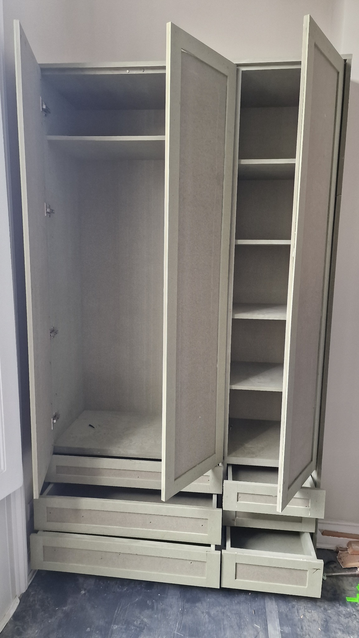 Otwarta, jasnoszara szafa z półkami i szufladami, widoczne zawiasy i niedoskonałości wykończenia.